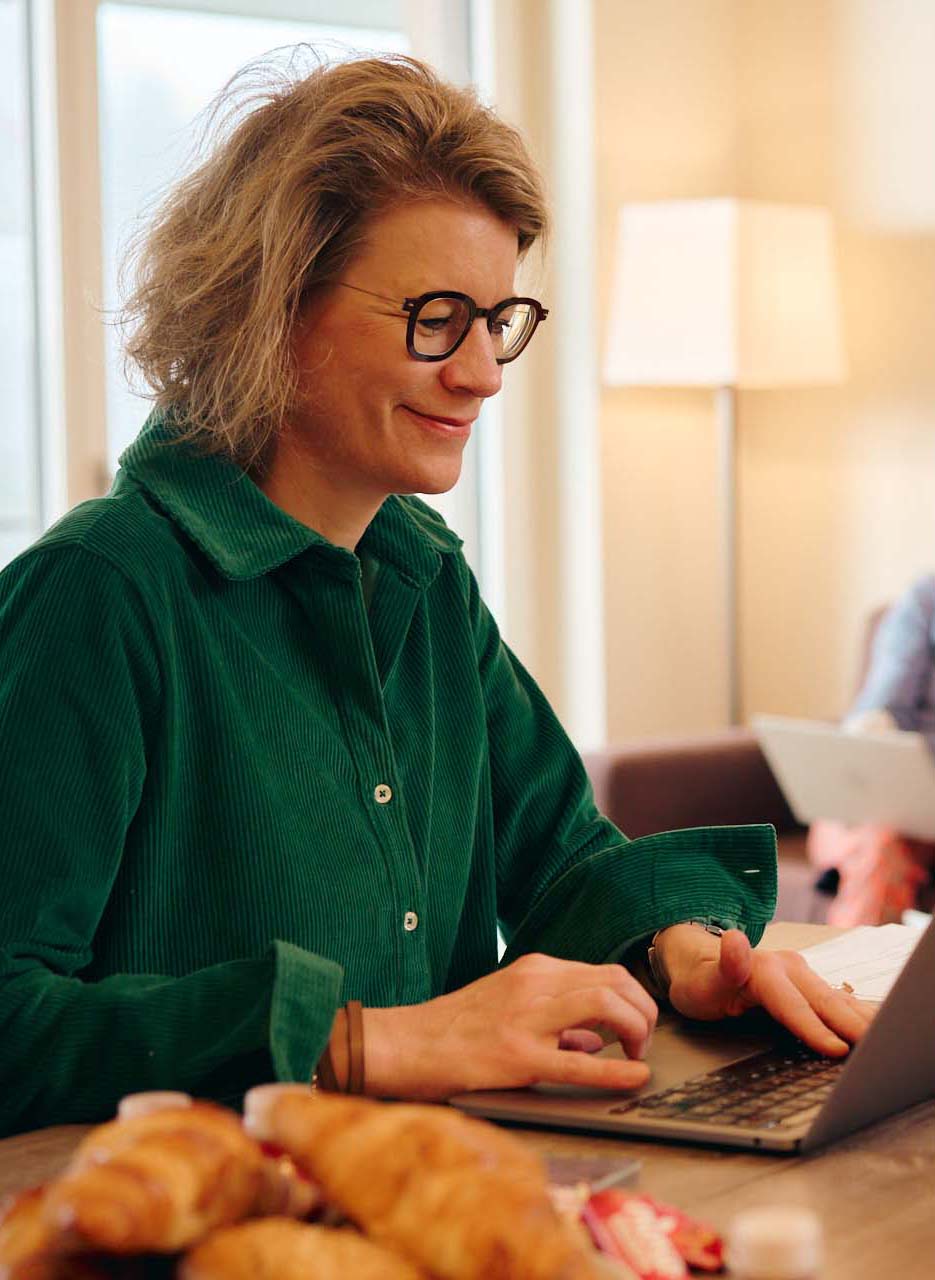 Bild 31 V8/Seite 12/Innovation: Nicoletta im Peaks Place während eines Workation-Aufenthaltes. Sie sitzt vor ihrem aufgeklappten Laptop und telefoniert mit einem Kunden. Auf dem Tisch stehen eine Teetasse und frische Brötchen. 