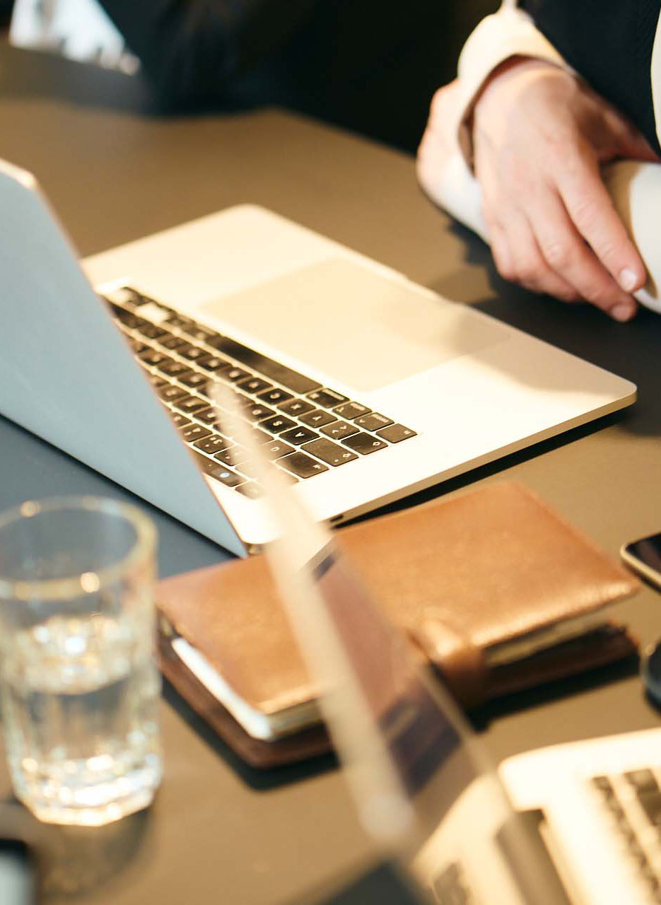 Blick auf einen Konferenztisch, auf dem zwei aufgeklappte Laptops, ein Wasserglas und ein heller Lederplaner stehen.
